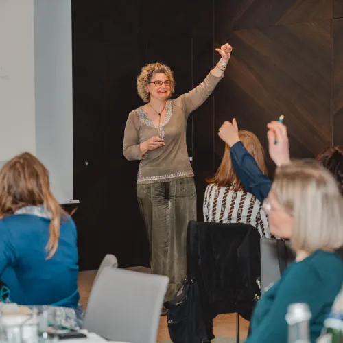 Frau spricht vor Publikum, einige Teilnehmer heben H&auml;nde.