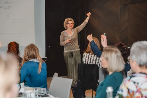 Frau spricht vor Publikum, einige Teilnehmer heben Hände.